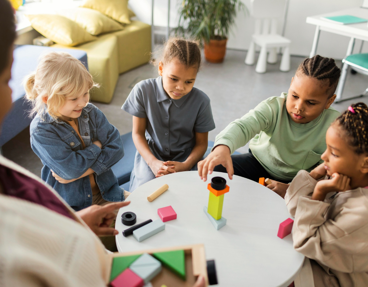 Four children sitting around a table playing with colorful wooden building blocks indoors.