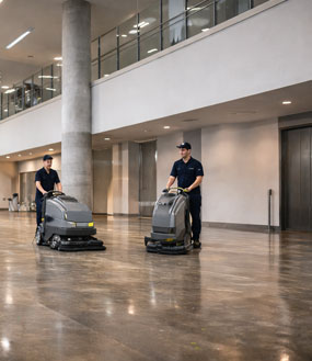 Two commercial cleaning professionals operating industrial floor scrubbers inside a spacious modern building with polished floors, high ceilings, and open interior architecture.