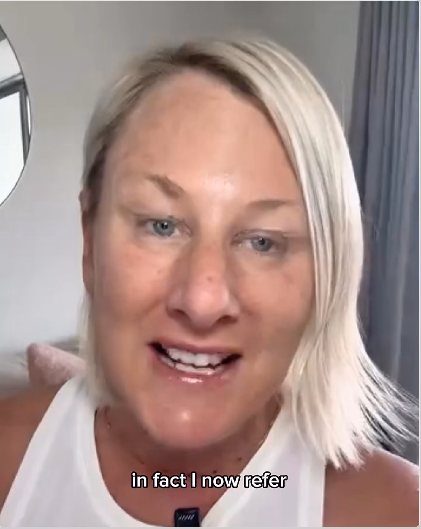 Close-up of a woman with short blonde hair speaking indoors, wearing a white top, with a round mirror and curtain visible in the background, and subtitle text that reads 'in fact I now refer'.