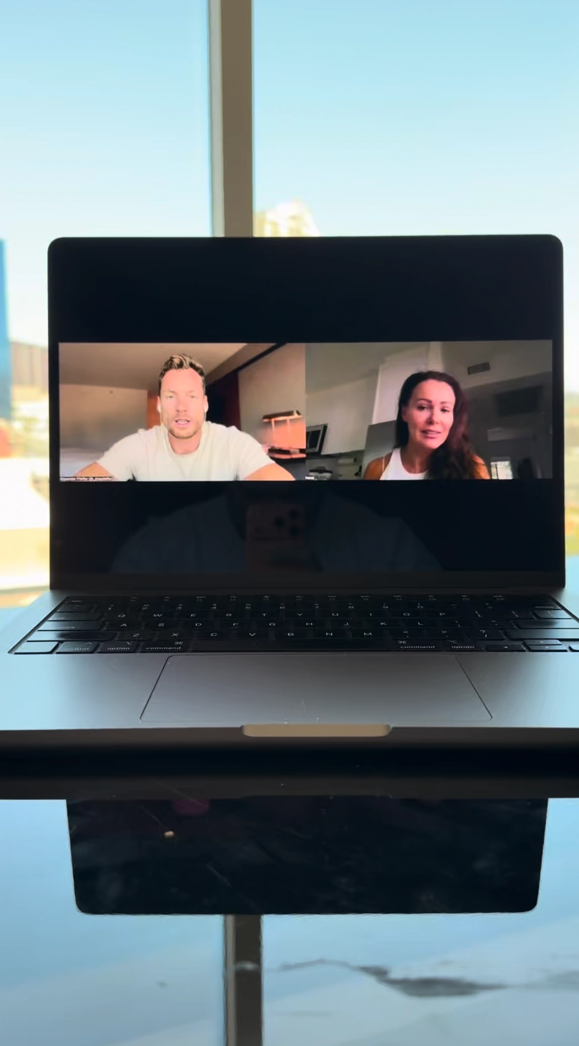 Laptop on a reflective surface showing a video call with a man in a white t-shirt and a woman with long dark hair.