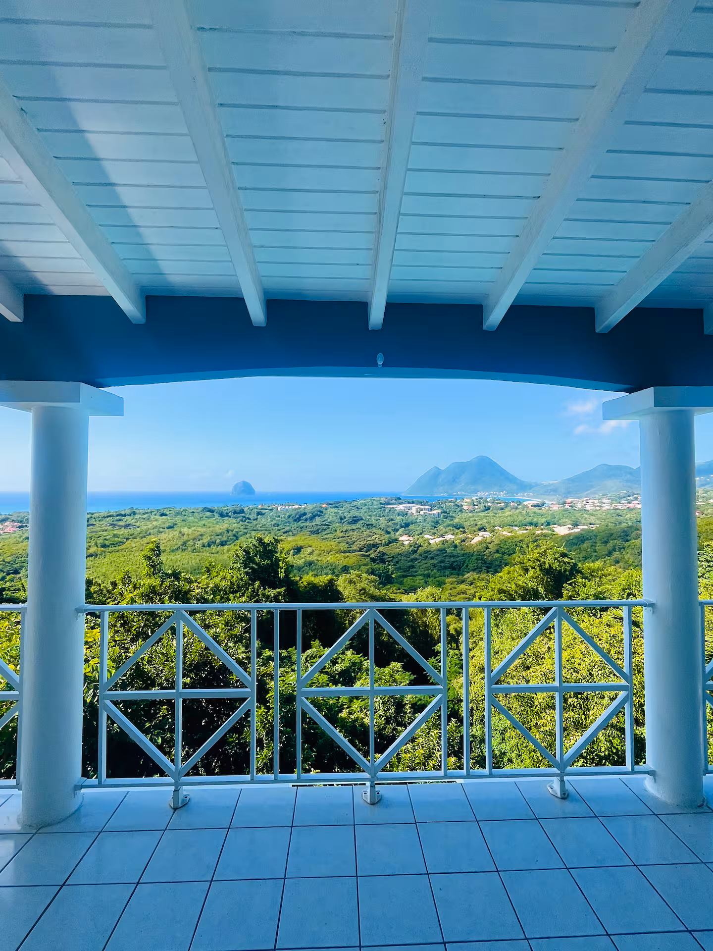 View of lush green hills and distant martinique mountains with the ocean on the horizon from a villa balcony with white railing and tiled floor. Luxury villa Martinique sea view Diamond Rock