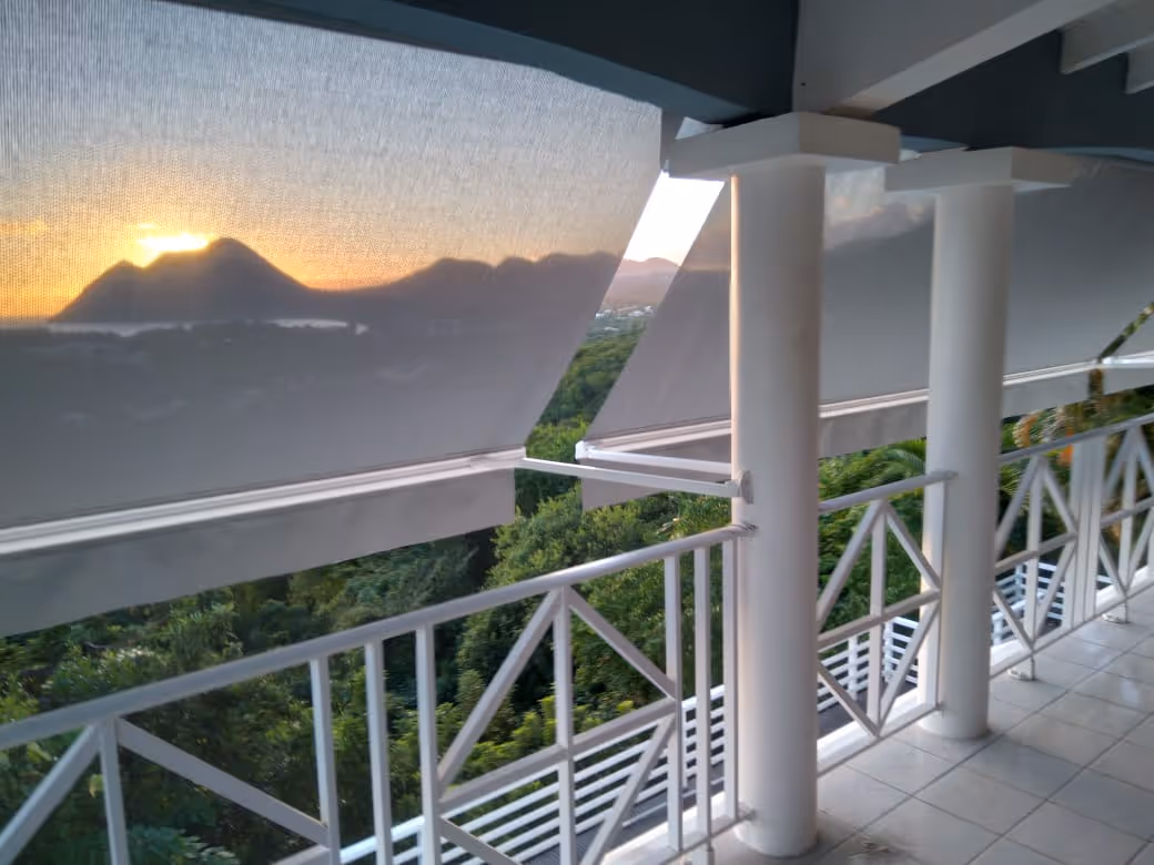 Sunset view of mountains and greenery seen through partially lowered outdoor shades on a balcony with white railings and columns. In a Martinique Diamant Villa for 20 Persons.