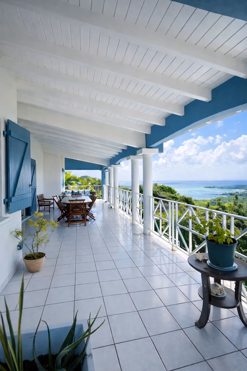Spacious balcony with white tiled floor, blue shutters, wooden dining set, potted plants, and a view of green foliage and ocean under a partly cloudy sky. In a Martinique Diamant Villa for 20 Persons.