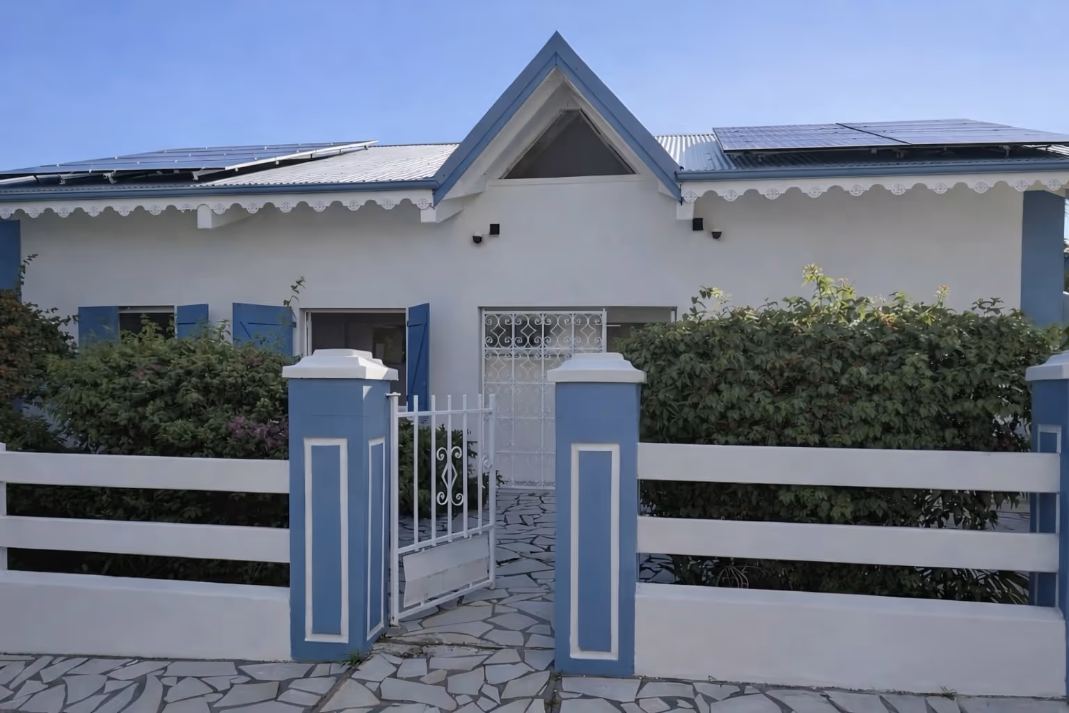 Small white house with blue shutters, blue and white fence, open gate, solar panels on the roof, and green bushes in front. In a Martinique Diamant Villa for 20 Persons.