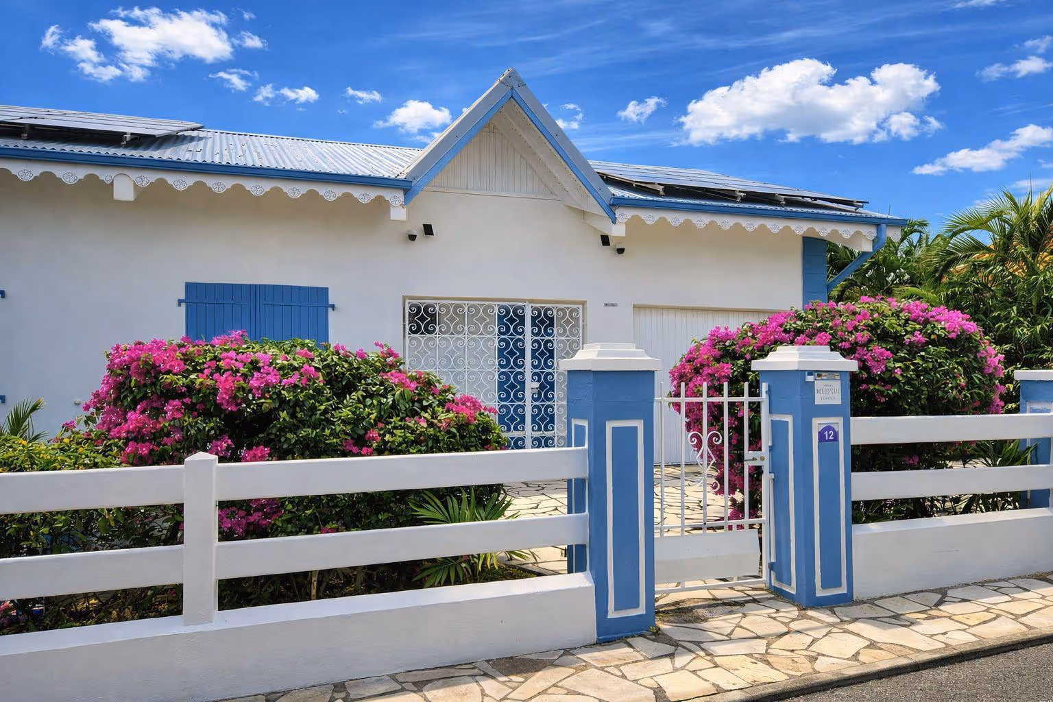 Small white house with blue shutters, blue and white fence, open gate, solar panels on the roof, and green bushes in front. In a Martinique Diamant Villa for 20 Persons.