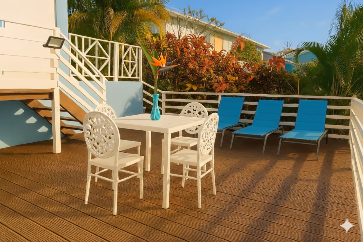Outdoor wooden deck with a white table and four ornate white chairs, a blue vase with a bird of paradise flower, and three blue lounge chairs against a white railing. In a Martinique Diamant Villa for 20 Persons.