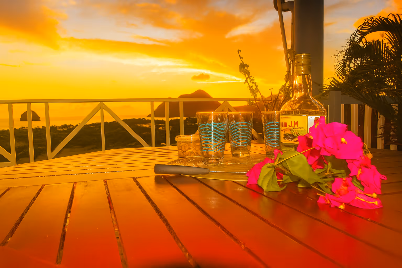 Wooden table on a balcony at sunset with a bottle, three decorated glasses, a container of nuts, and bright pink flowers. In a Martinique Diamant Villa for 20 Persons.