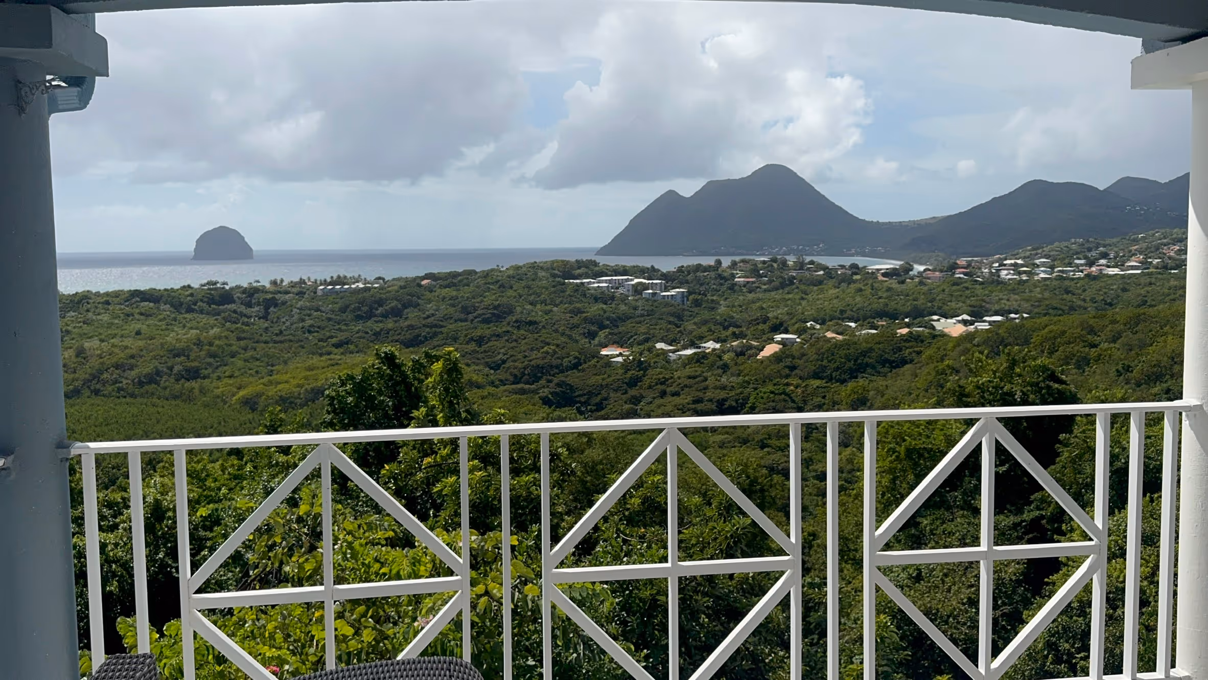 View from balcony with white railing overlooking dense green forest, distant mountains, and a small island in the ocean under a cloudy sky. In a Martinique Diamant Villa for 20 Persons.