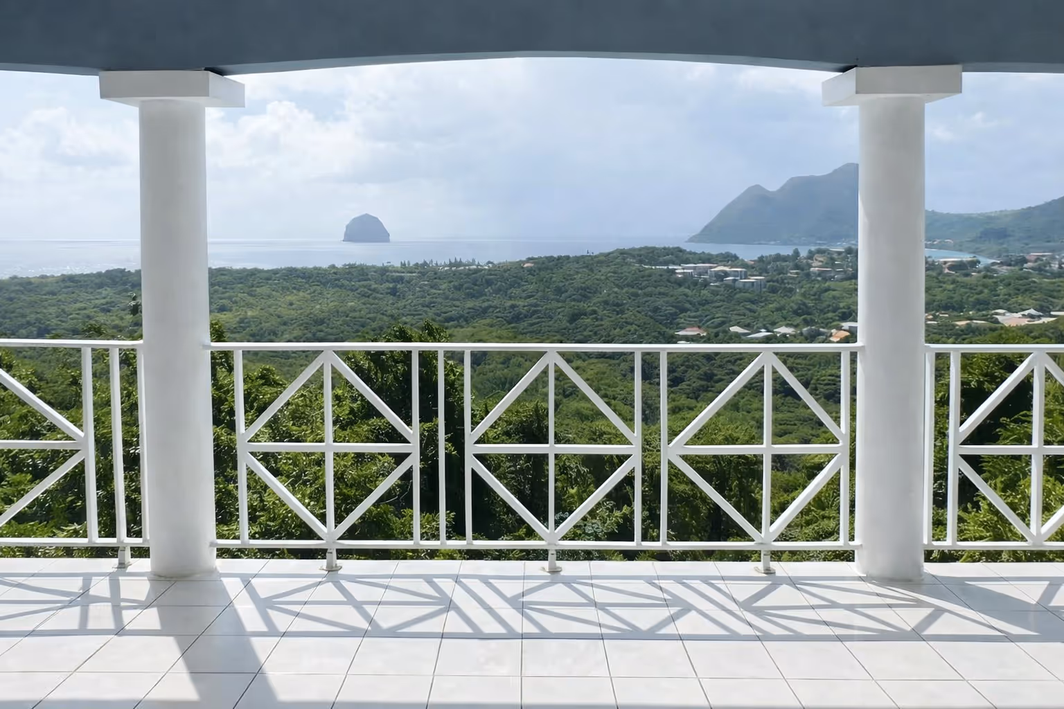Ocean view from a white-tiled balcony with white railing and pillars, overlooking green hills and distant mountains under a cloudy sky. In a Martinique Diamant Villa for 20 Persons.