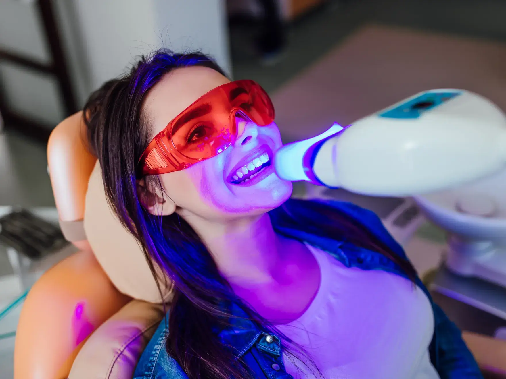 Woman wearing protective red glasses receiving blue light teeth whitening treatment while reclining in dental chair.