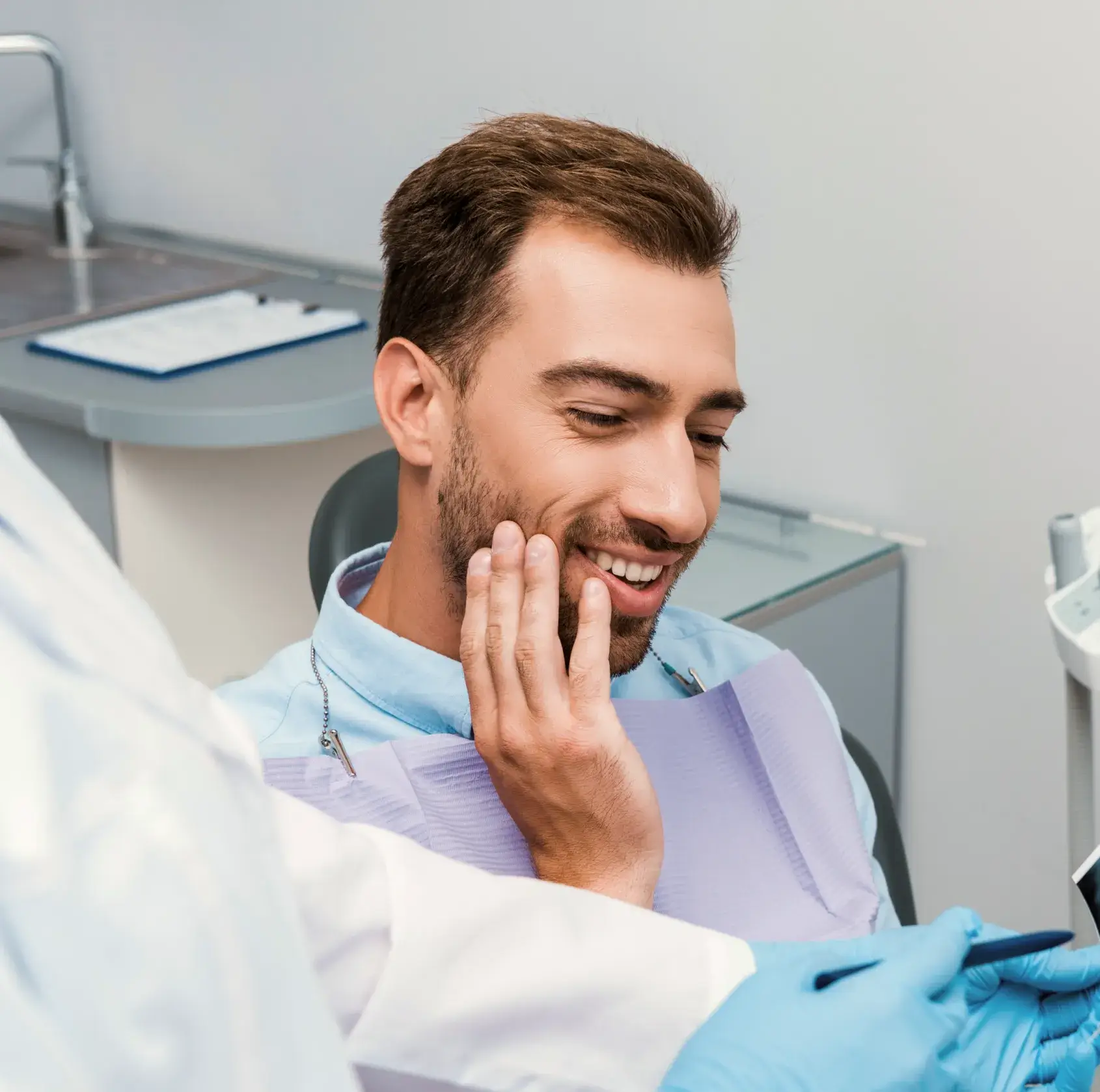 Smiling man in dentist chair holding his cheek while a dentist wearing blue gloves shows him an X-ray.
