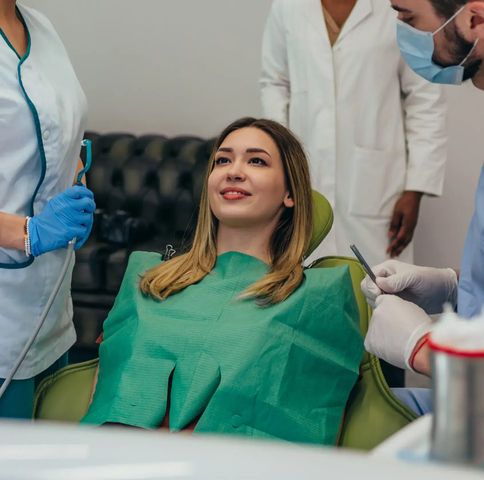 Smiling woman sitting in a dental chair with a green bib, surrounded by two dental professionals preparing tools.