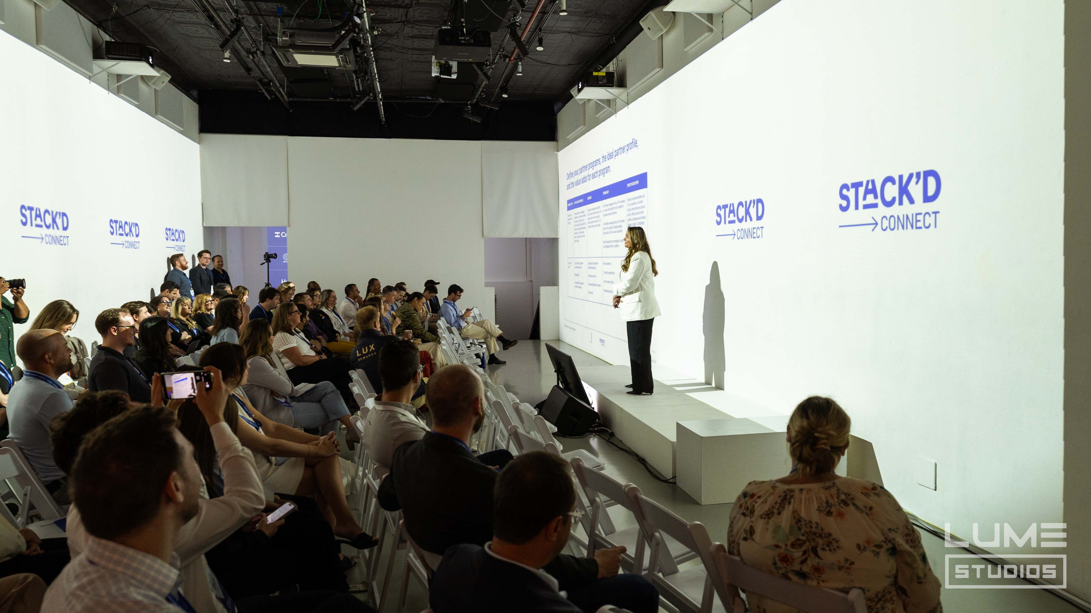 Speaker presenting to a seated audience under Stack'd Connect projections at LUME Studios