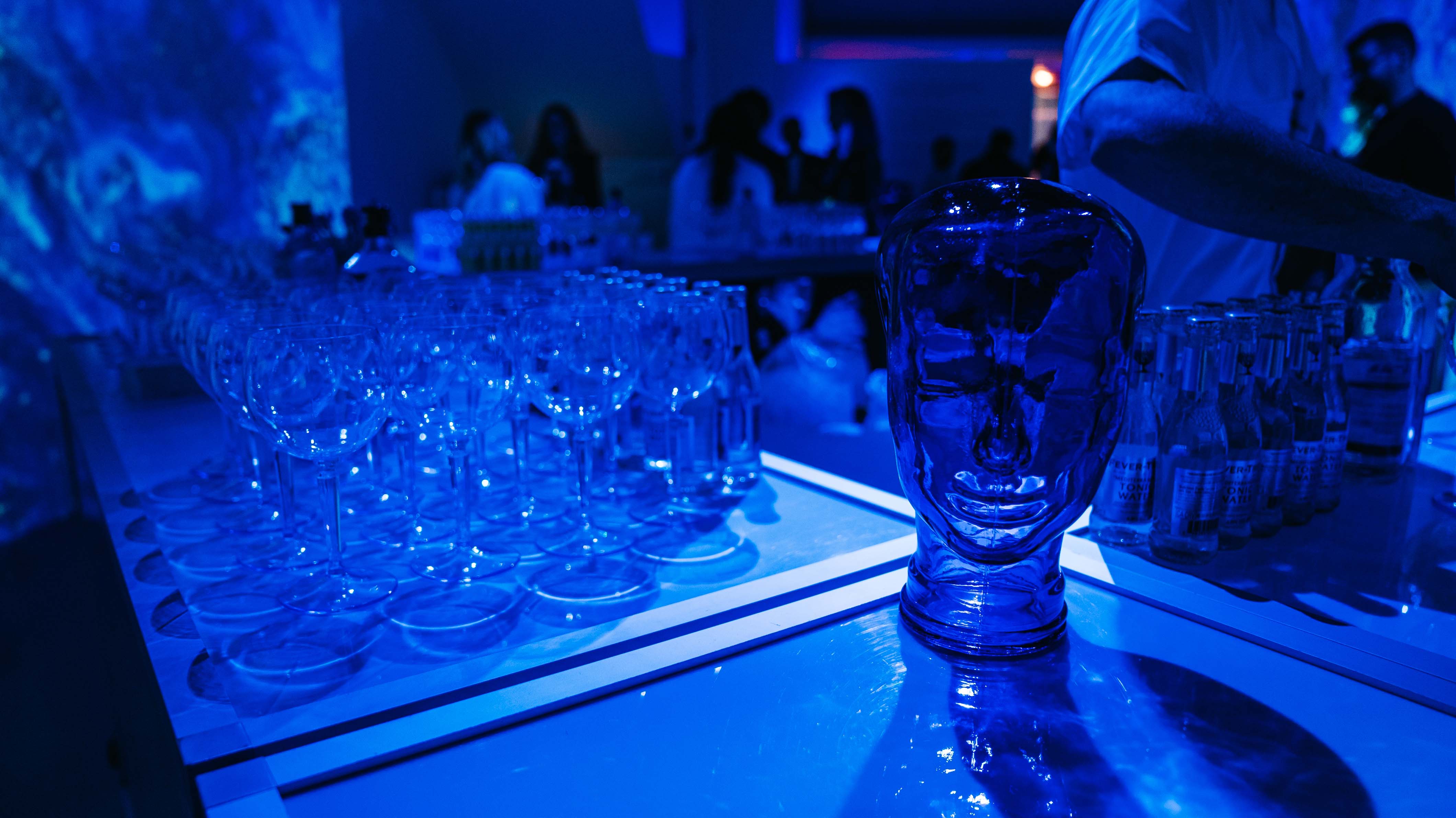 Blue-lit bar with rows of glasses and a glass head sculpture at the Bombay Sapphire x Dan Lam event