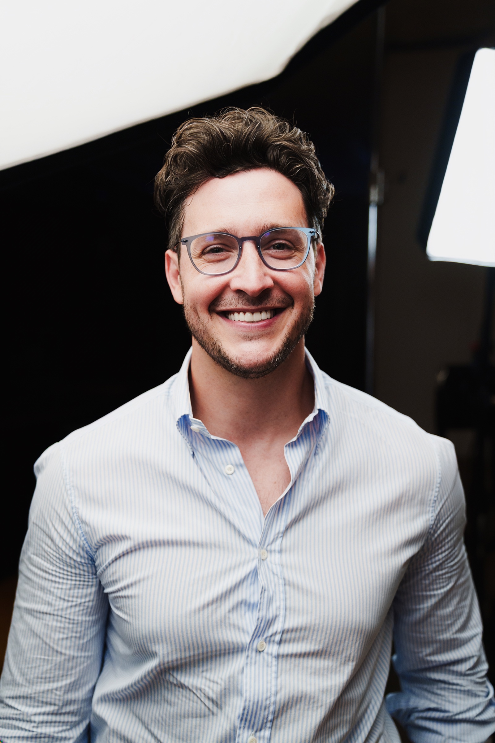Doctor Mike in a pale blue shirt and glasses smiles beside a softbox on the podcast set.
