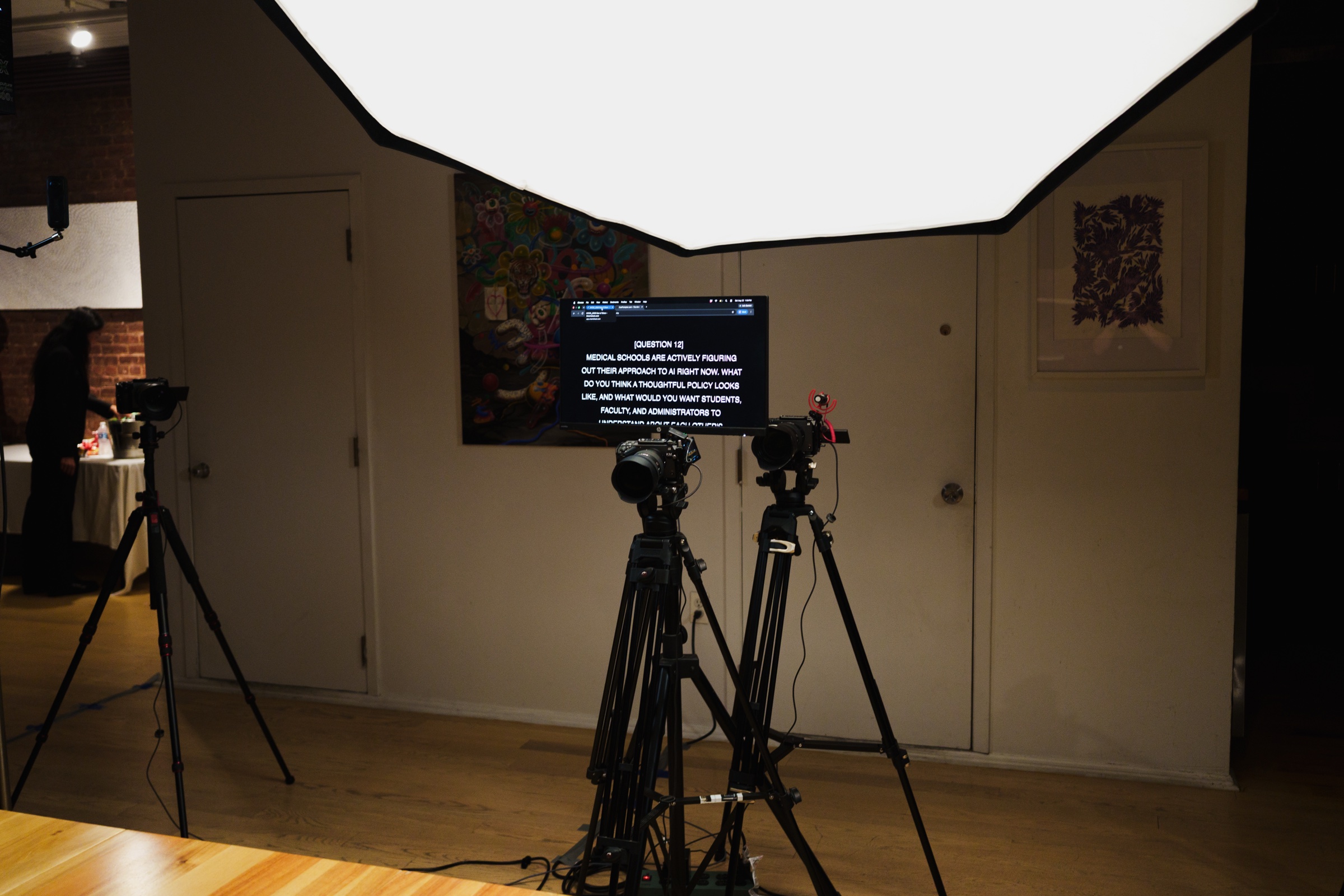 A monitor displays a discussion question behind two cameras under a large overhead softbox.