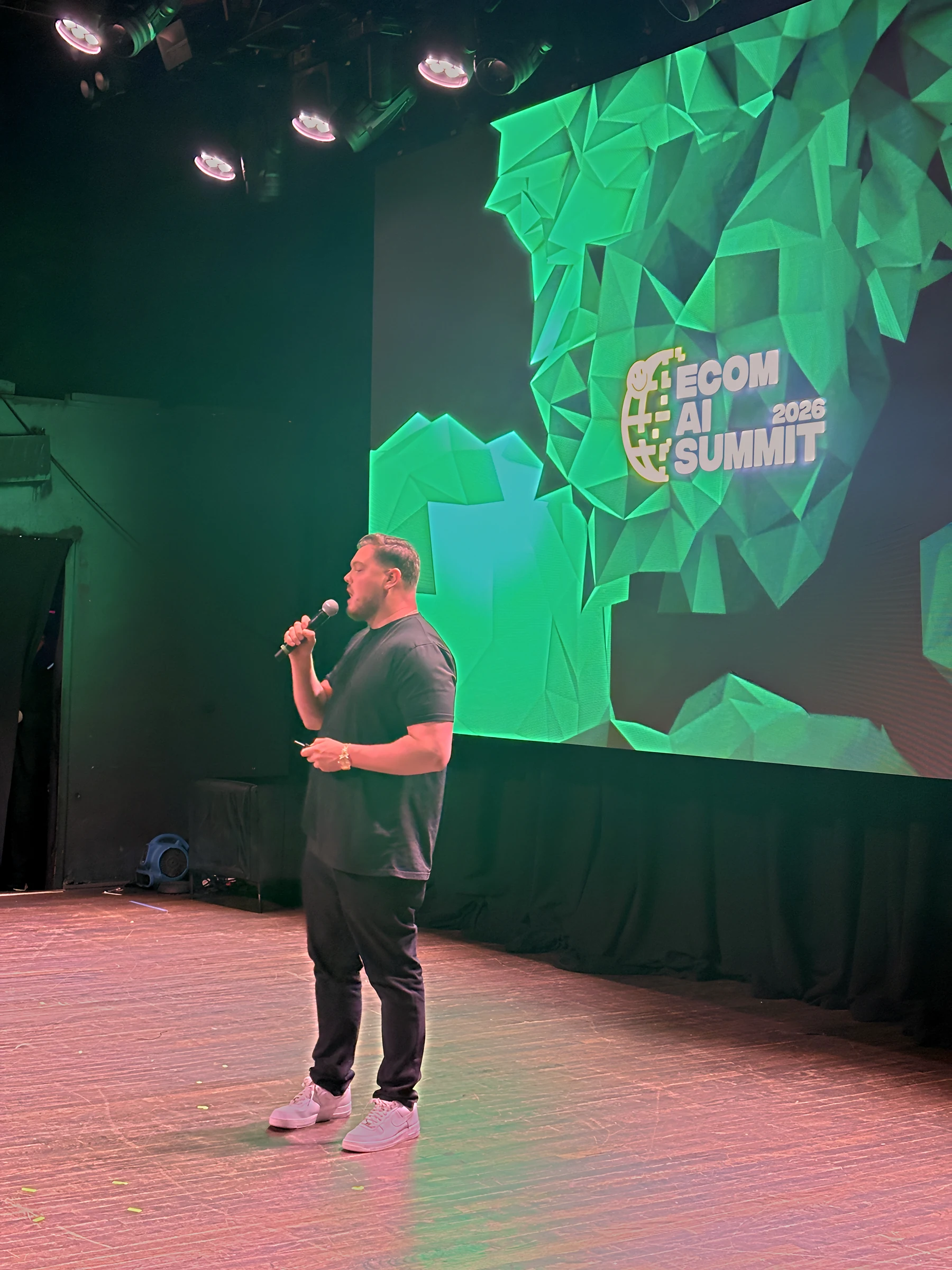 A keynote speaker on the Webster Hall stage in front of the green Ecom AI Summit 2026 graphic on the LED wall.