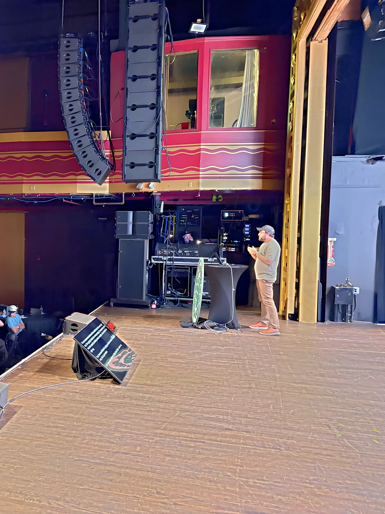 A presenter speaks at the edge of the Webster Hall stage under the line array speakers, with the show rack behind.