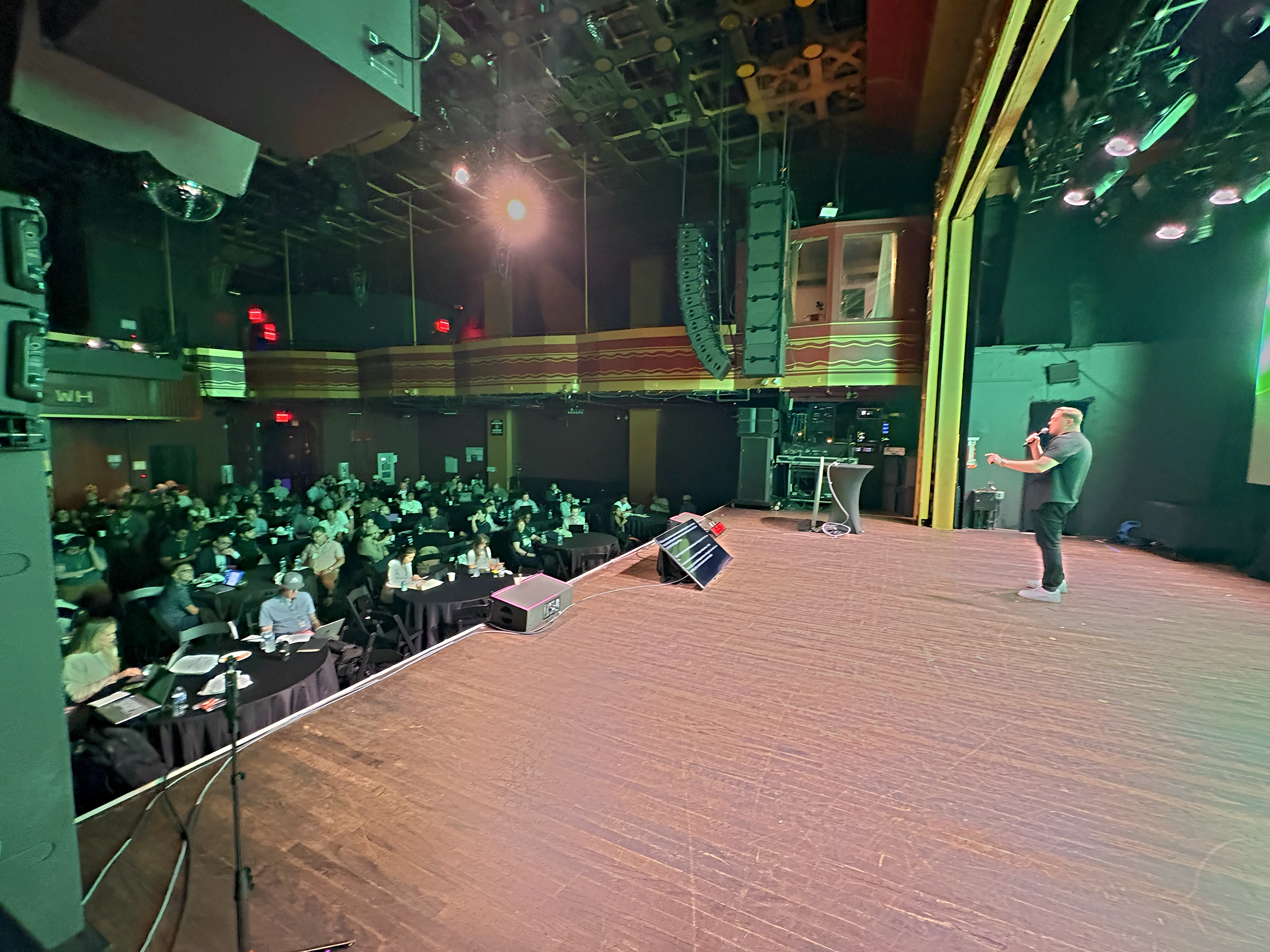 View from the stage wing of a speaker addressing the Ecom AI Summit audience under green stage lighting.