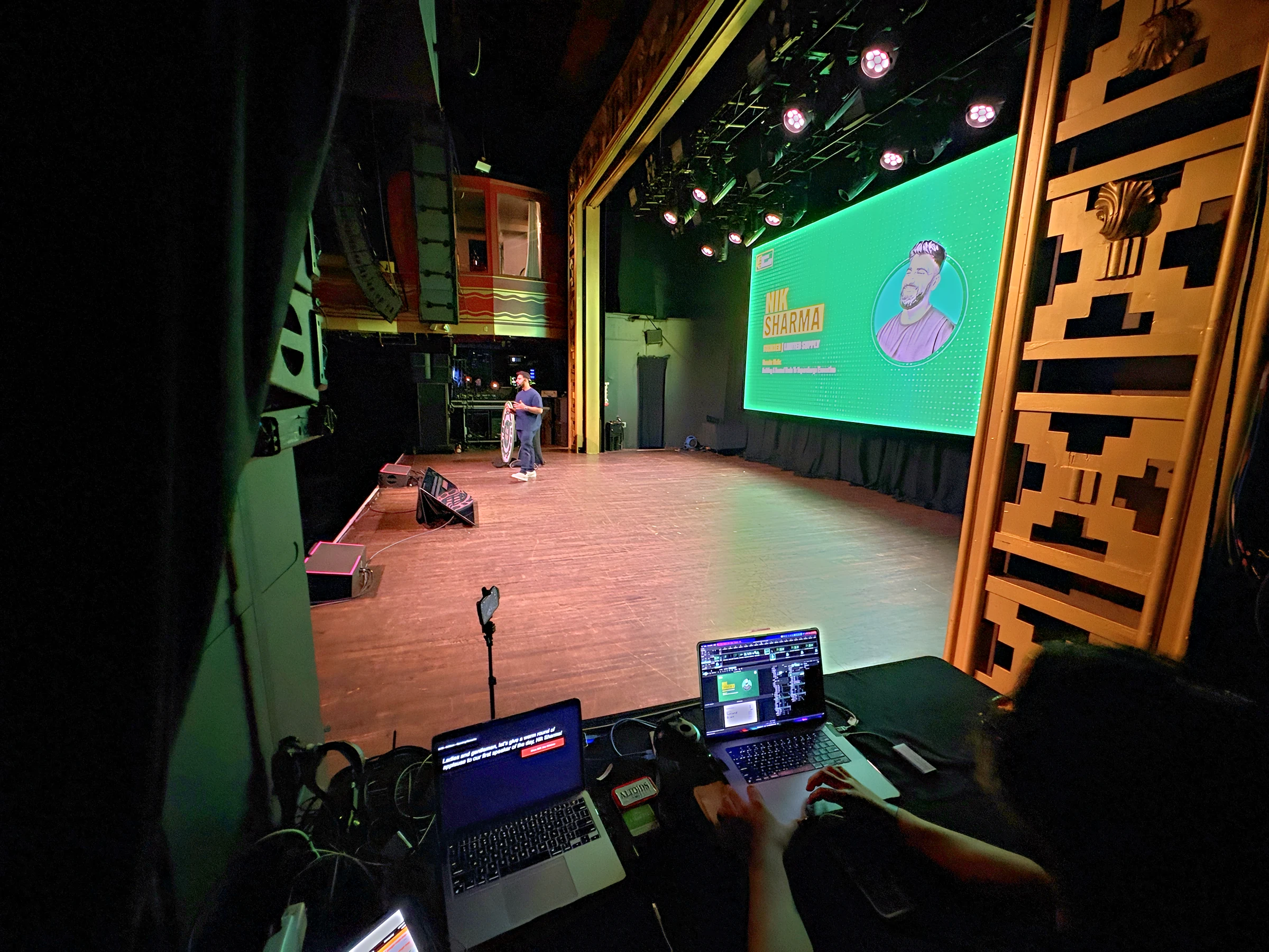 Show control laptops at the side-stage booth with a speaker title card on the green LED wall at Webster Hall.