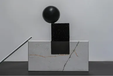 Black sphere balanced on a small black rectangular block above a larger white marble block with visible veins.