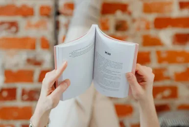 Person holding an open book with visible text, sitting in front of a blurred brick wall.