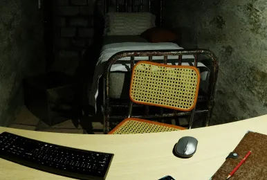 Small room with an old metal bed, a cane chair, and a desk with a keyboard, mouse, and pencil.