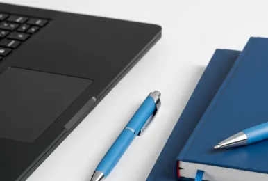 Black laptop keyboard partially visible next to two blue notebooks and two blue pens on a white surface.