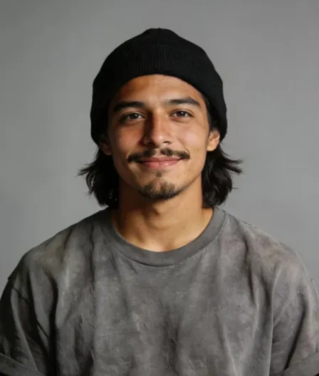Young man with medium-length dark hair wearing a black beanie and gray sweatshirt smiling against a plain gray background.
