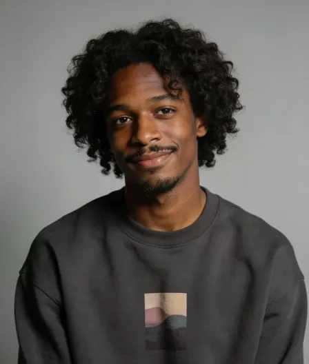 Young man with curly hair, wearing a dark sweatshirt, smiling against a plain gray background.
