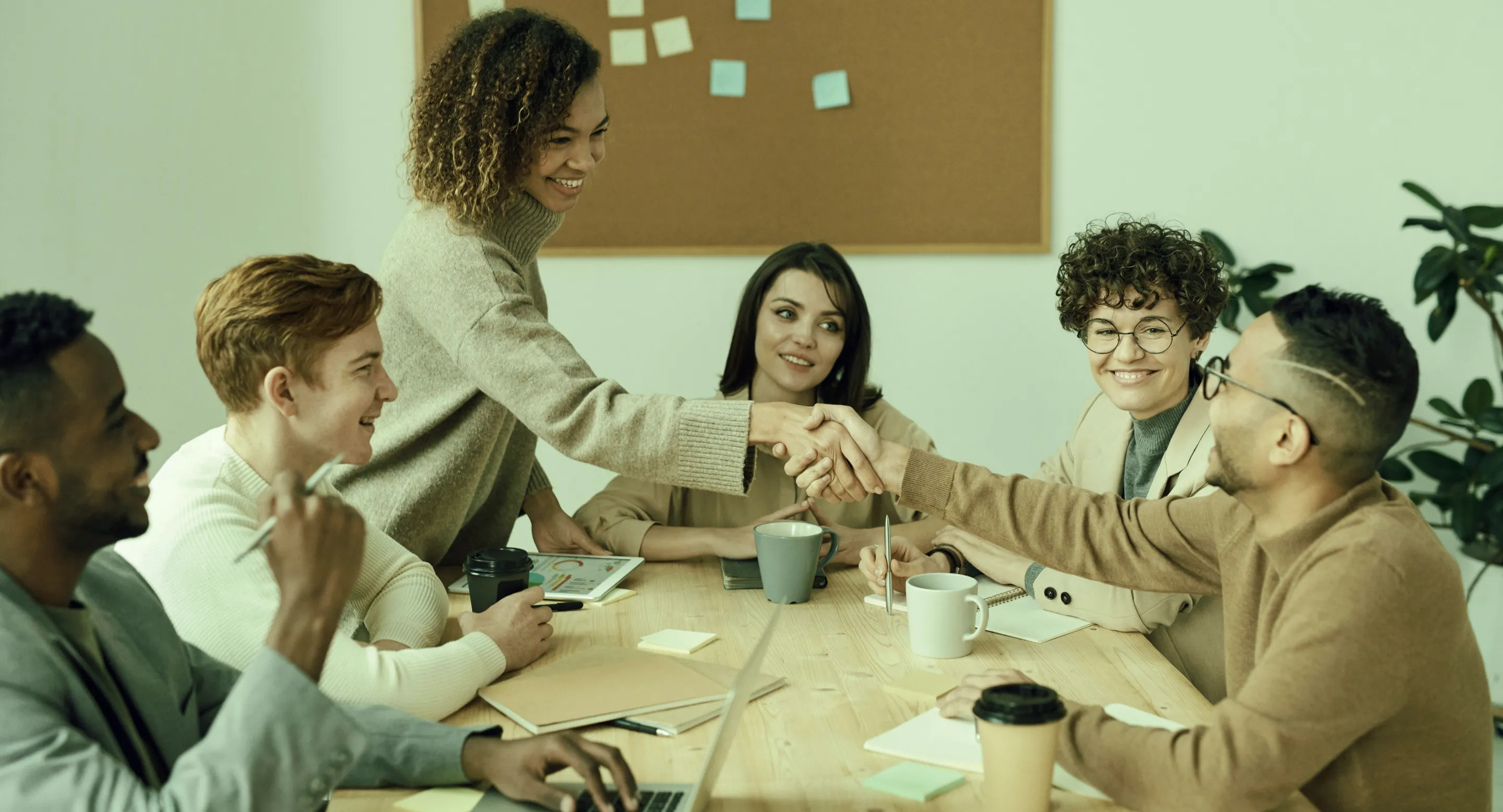 A diverse group of five people around a table in a meeting, two are smiling and shaking hands.