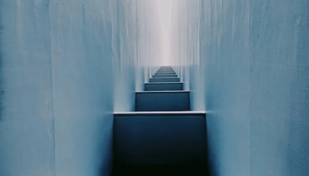 Narrow blue staircase between two tall blue walls leading to a bright light at the top.