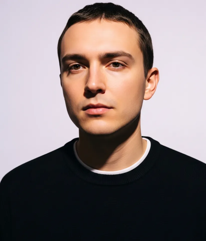 Young man with short dark hair wearing a black sweater with a white collar against a plain light background.