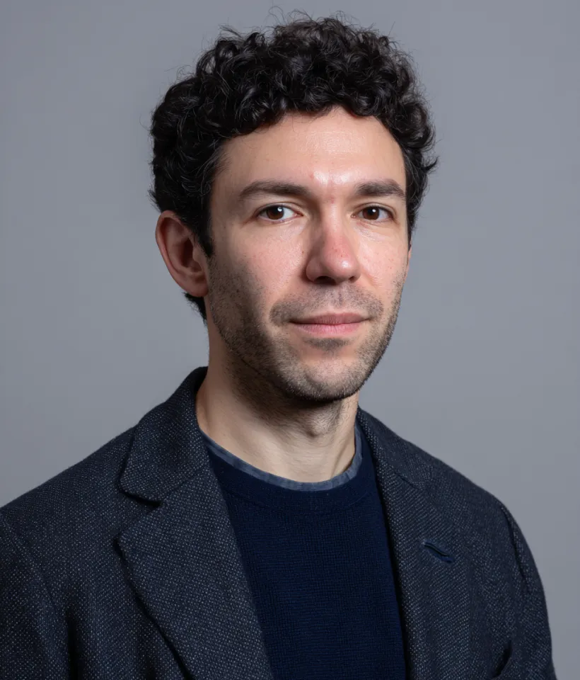 Man with curly dark hair wearing a dark blazer and navy sweater against a gray background.