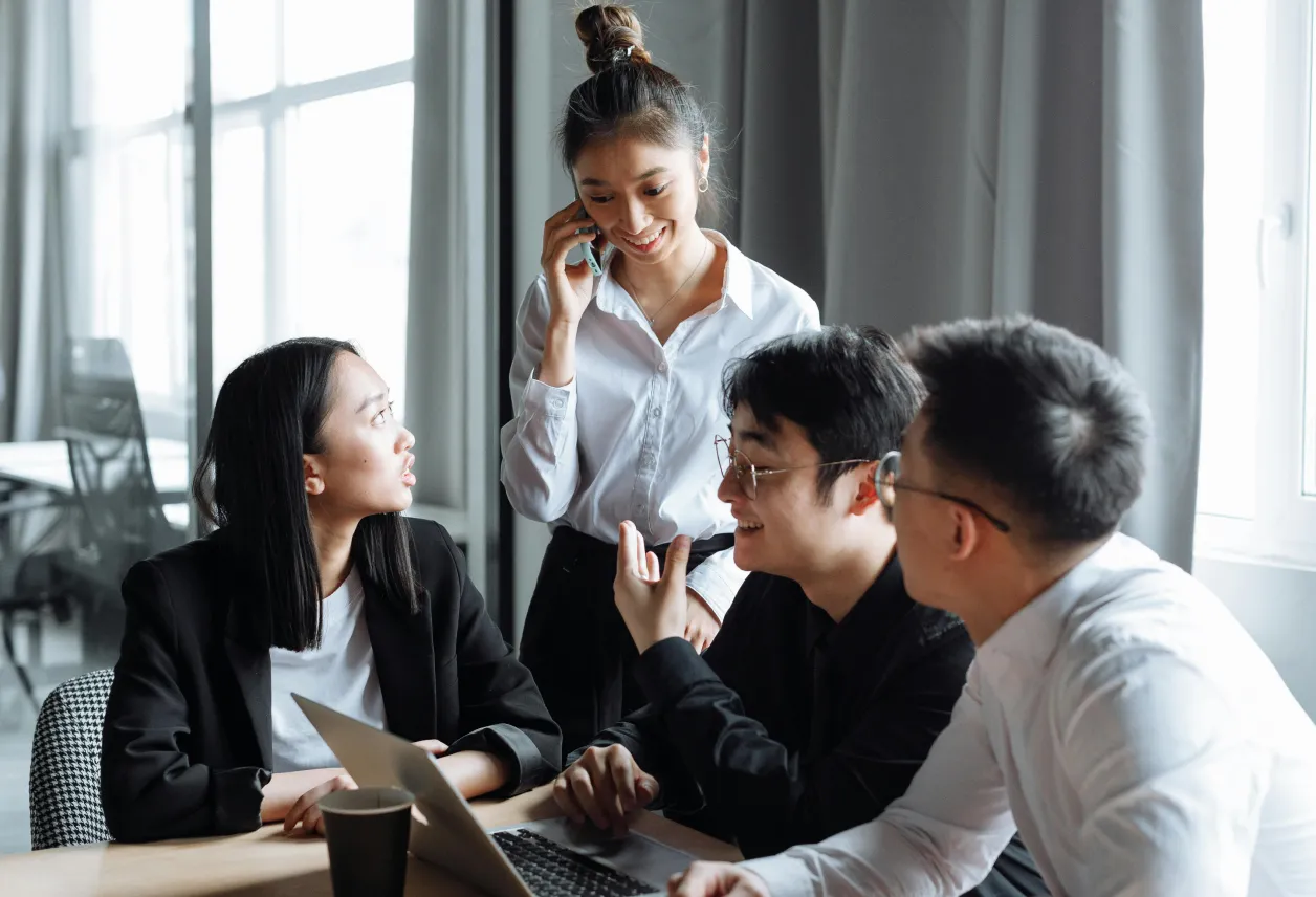 Four young professionals having a discussion in an office, one woman standing while talking on the phone and three colleagues seated with a laptop.
