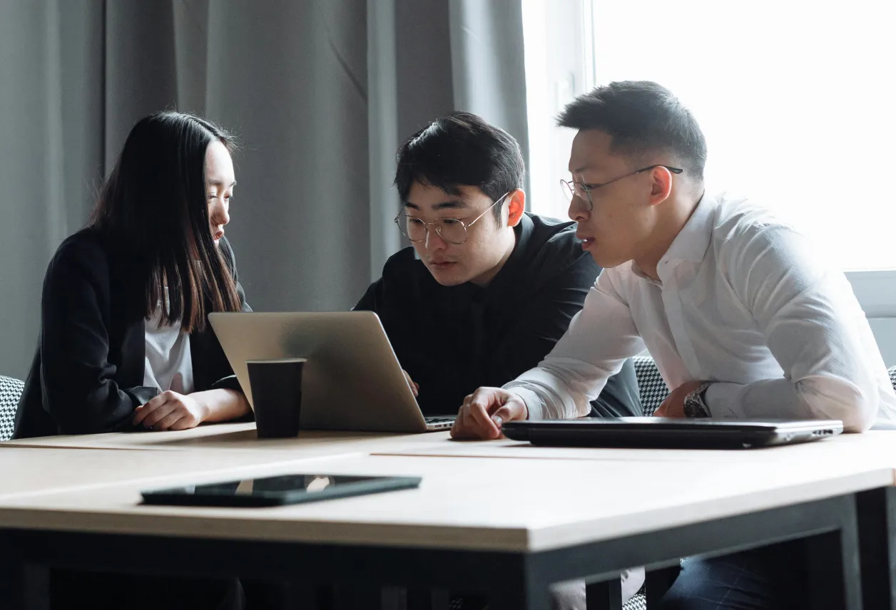 Three young professionals focused on a laptop during a meeting at a light wooden conference table.