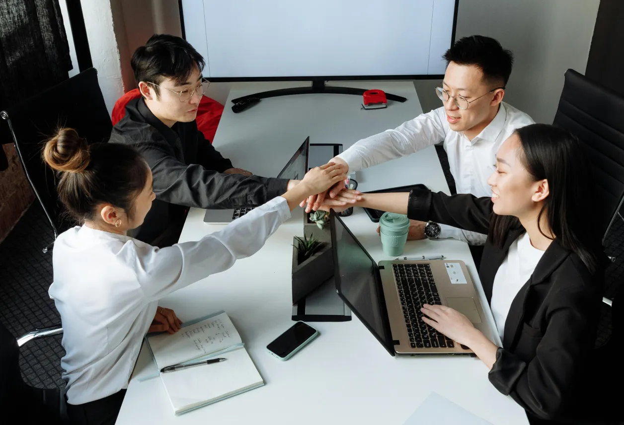 Four coworkers sitting around a table joining hands in a team gesture during a meeting with laptops and notebooks.
