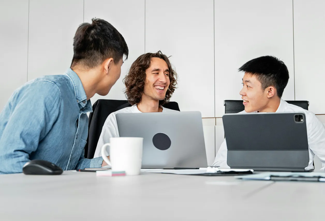 Three young men sitting around a table with laptops and a coffee mug, engaged in a lively discussion.