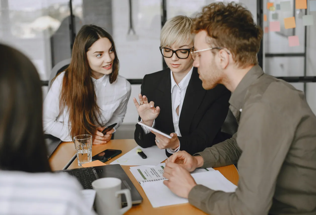 Group of four young professionals discussing work around a table with documents, a laptop, and smartphones in a modern office.