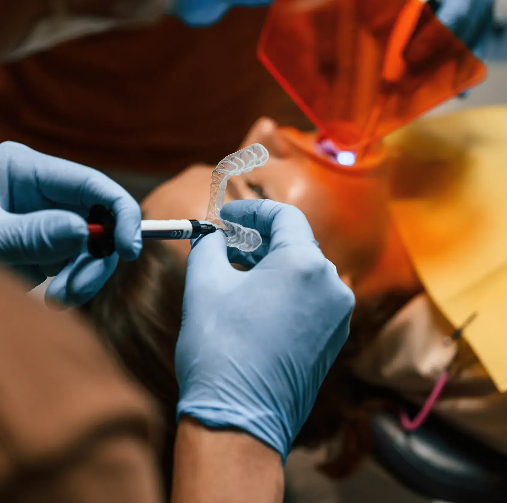 Gloved hands holding a dental aligner while using a marker with a patient lying down in the background with protective orange eyewear.