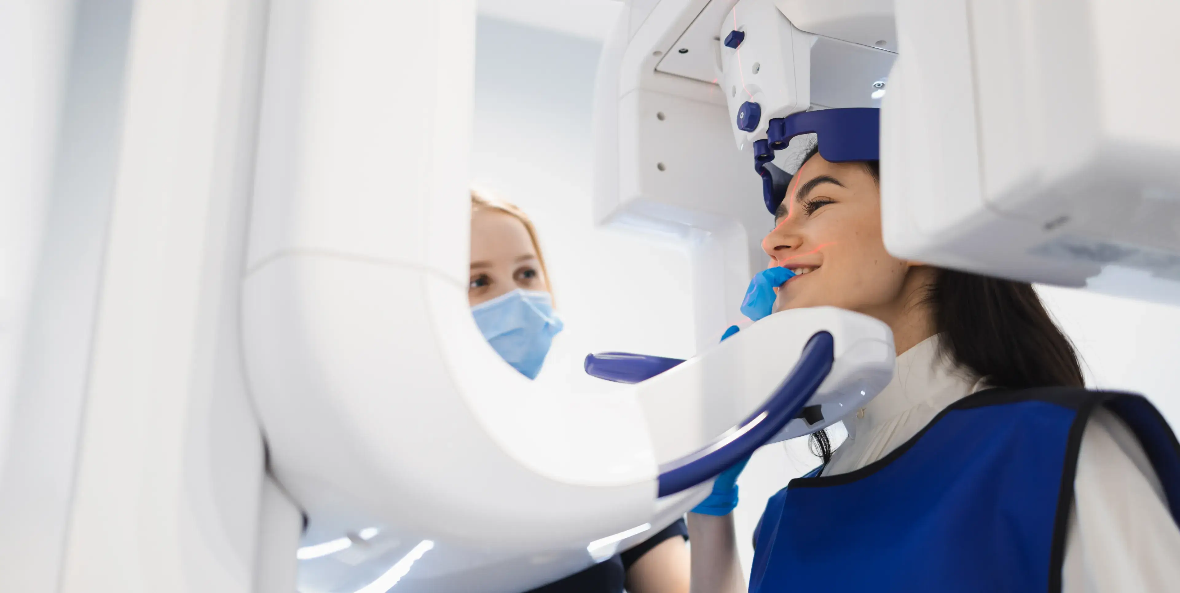 Woman wearing blue protective vest undergoing a dental X-ray while a masked technician assists.