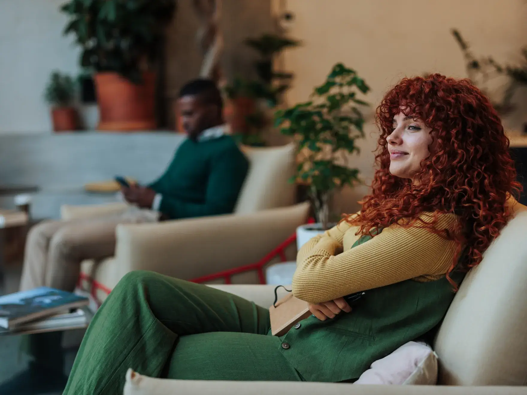 Smiling woman with curly red hair sitting relaxed on a beige armchair holding a notebook, with a man blurred in the background using a phone.