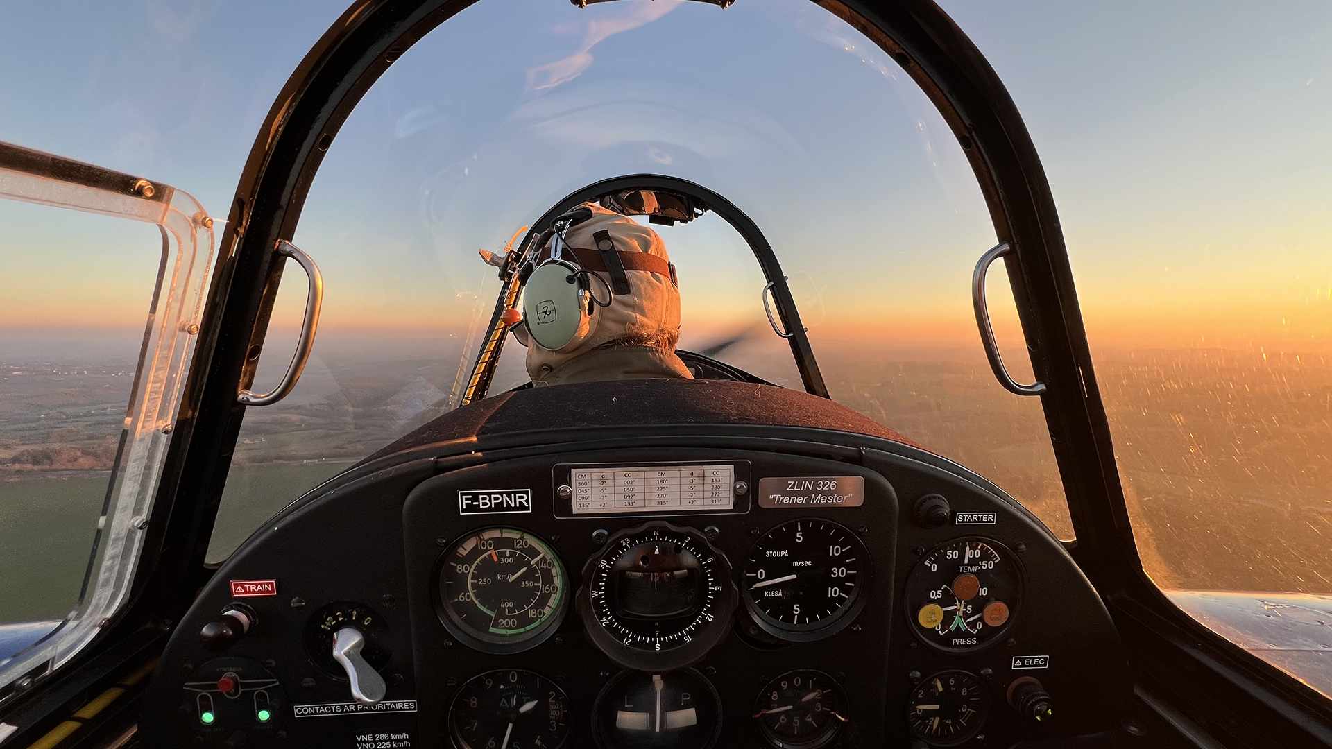 Pilot wearing headphones flying a ZLIN 326 Trainer Master airplane at sunset, viewed from behind inside the cockpit.