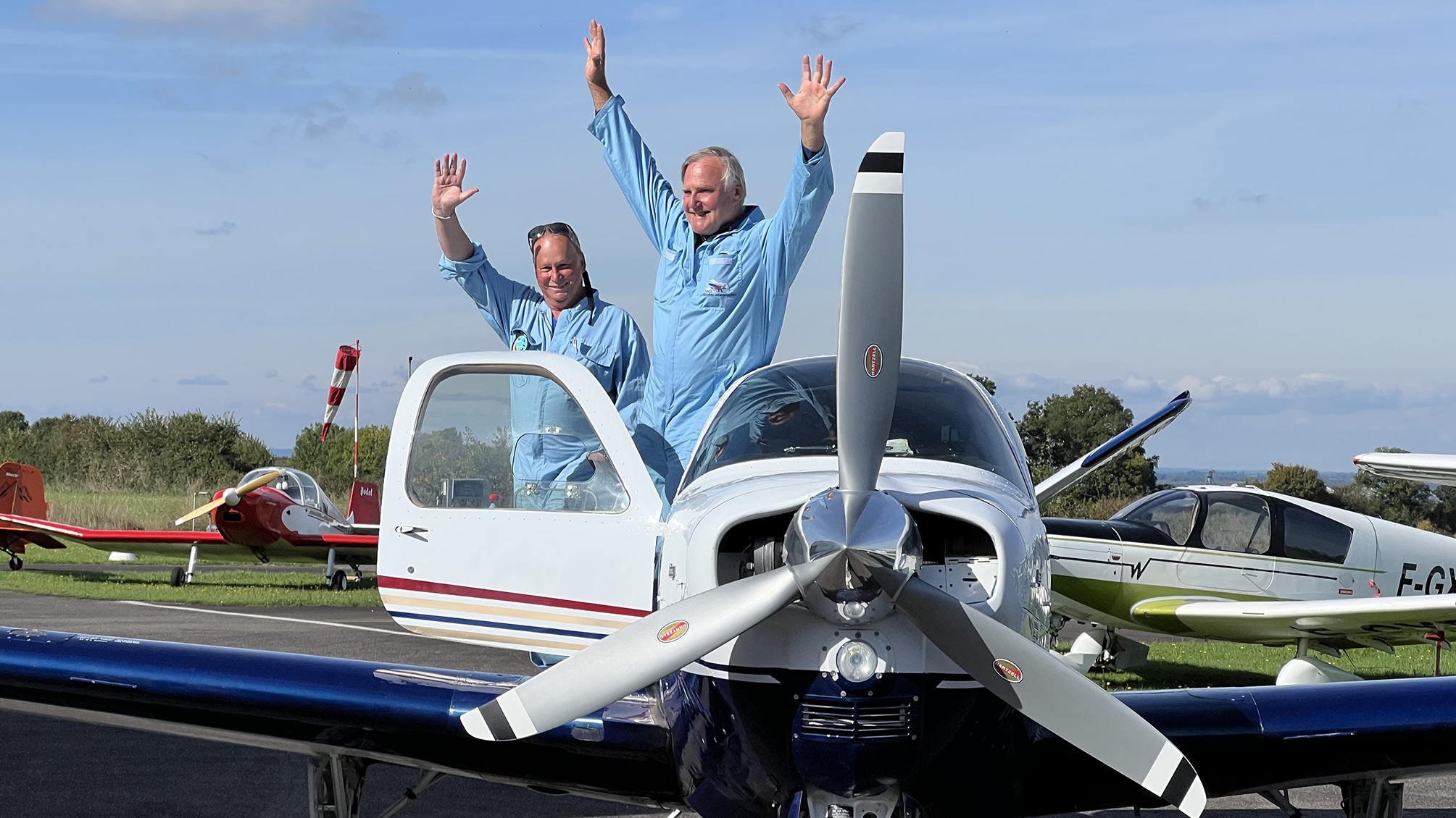 Two men in blue flight suits standing on a small white and blue propeller airplane with arms raised in celebration at an airfield.