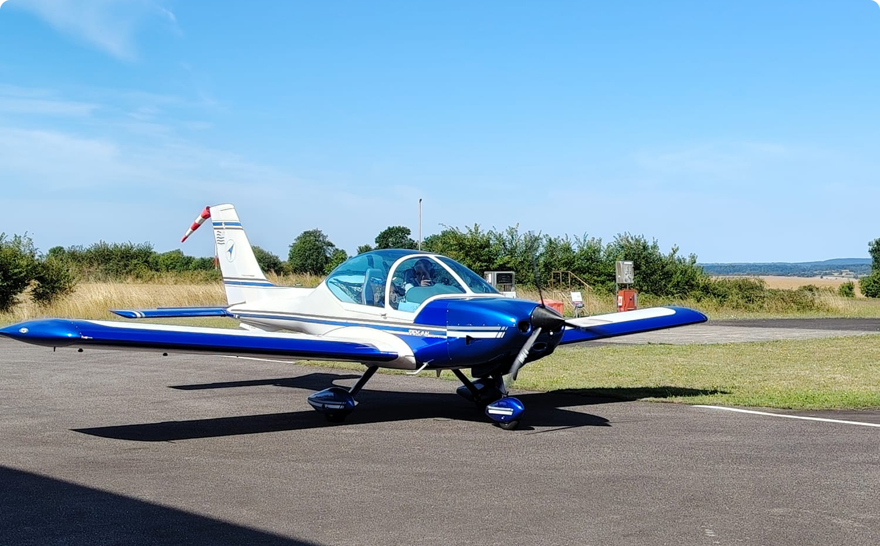 Small white and blue single-engine airplane parked on a tarmac under a clear blue sky.