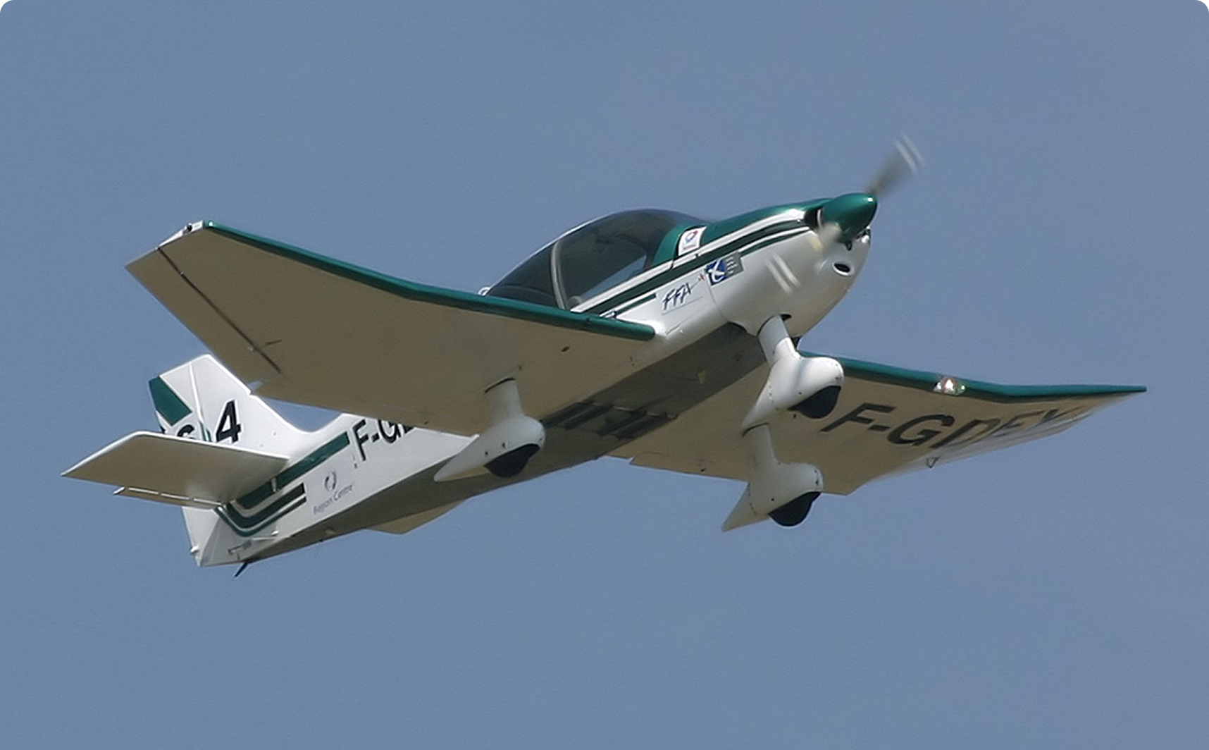 White and green single-engine light airplane flying against a clear blue sky.