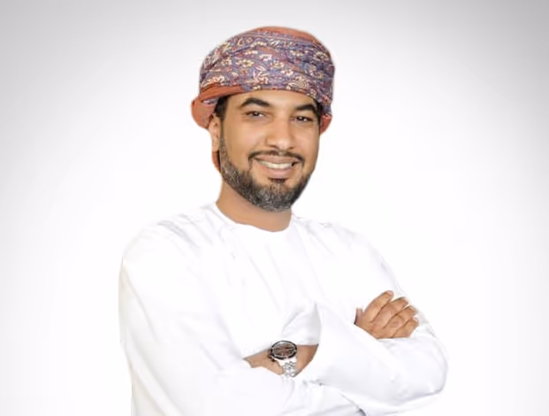 Smiling young woman with curly brown hair and glasses wearing a light sweater and necklace against a plain background.