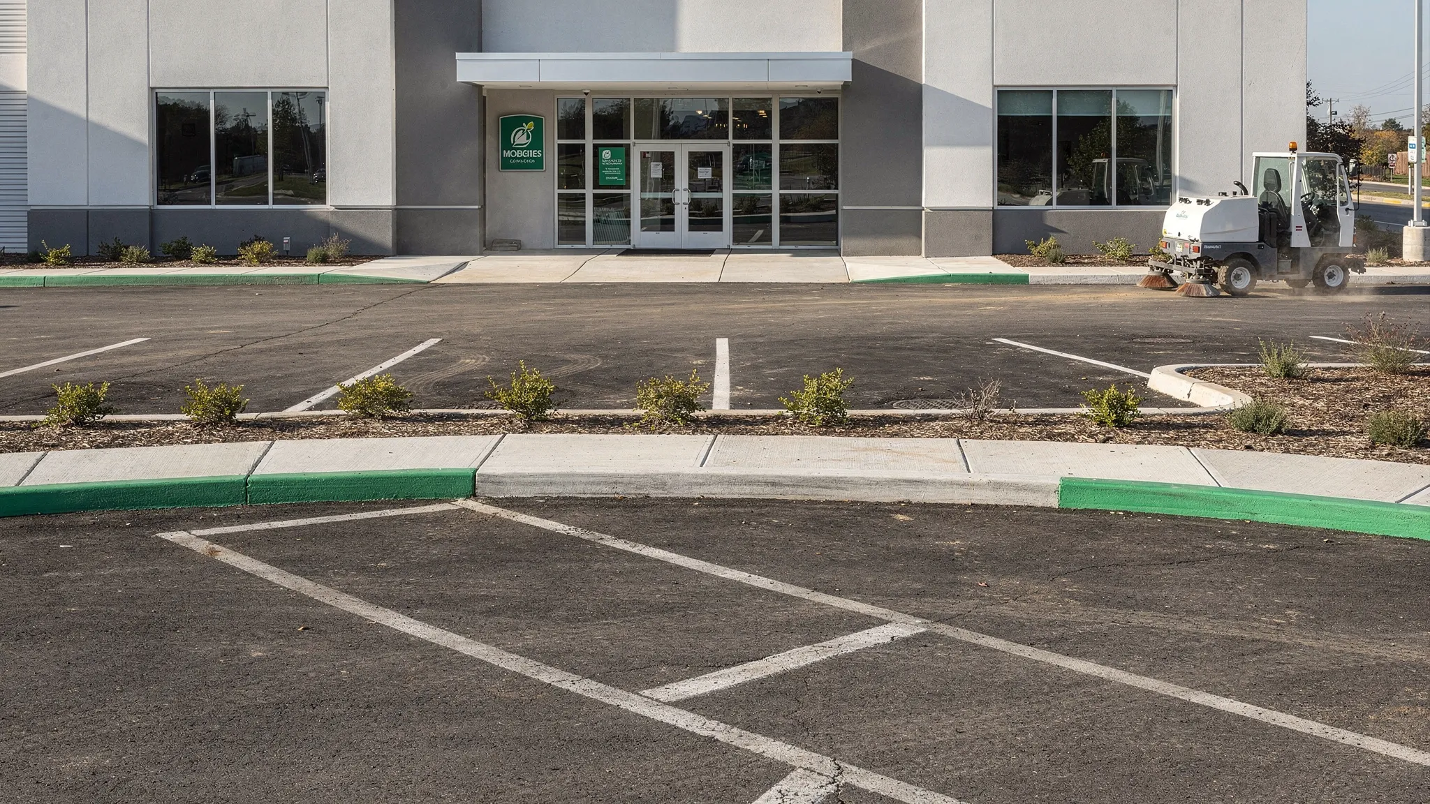 A clean, finished construction site exterior with freshly swept parking lot, clear curb lines, clean sidewalks at the main entrance, and a street sweeper working near the curb in the background.