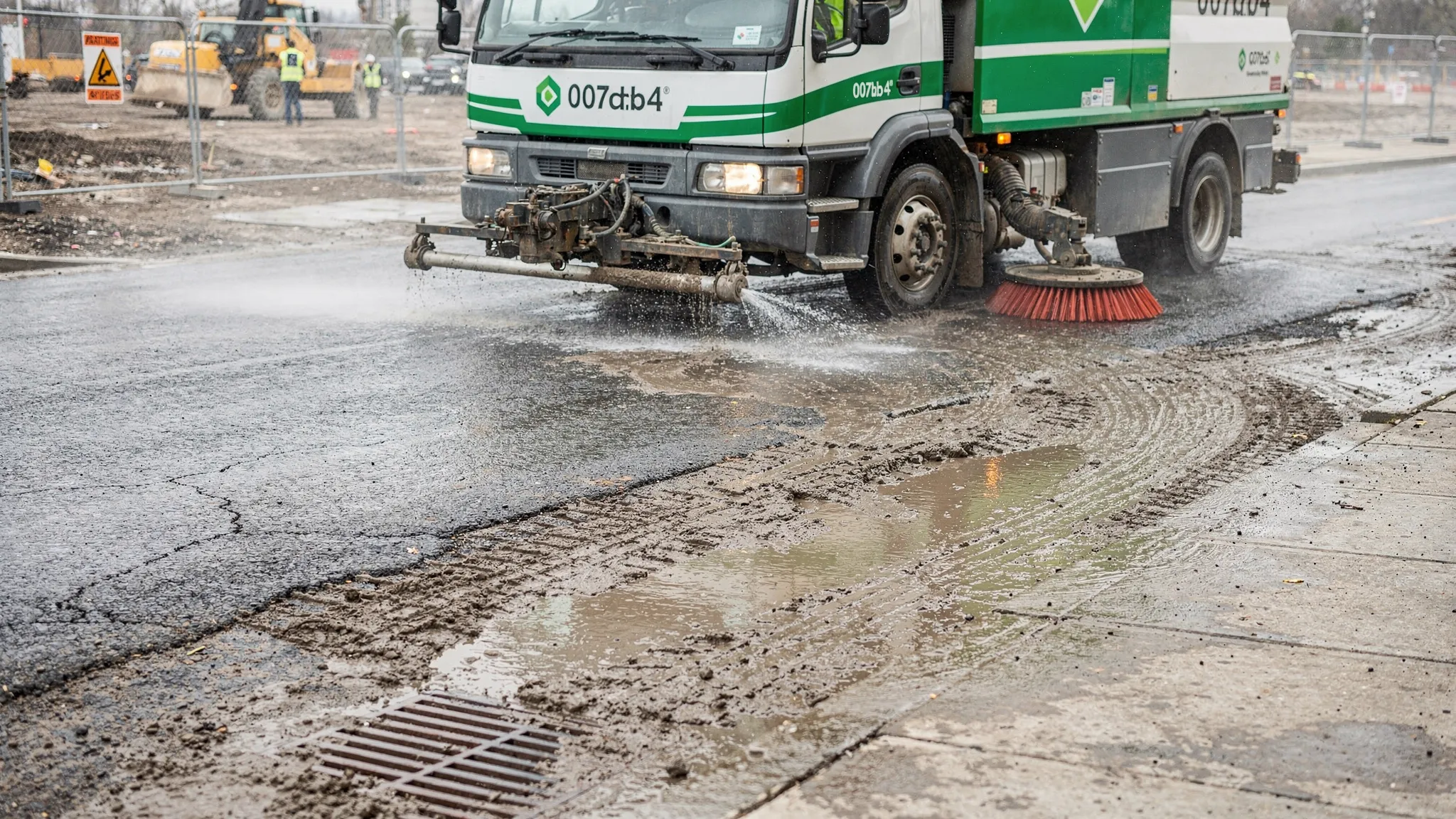 A professional street sweeper cleaning a muddy construction entrance and adjacent roadway, with visible mud tracking lines being removed from asphalt near a curb and storm drain.