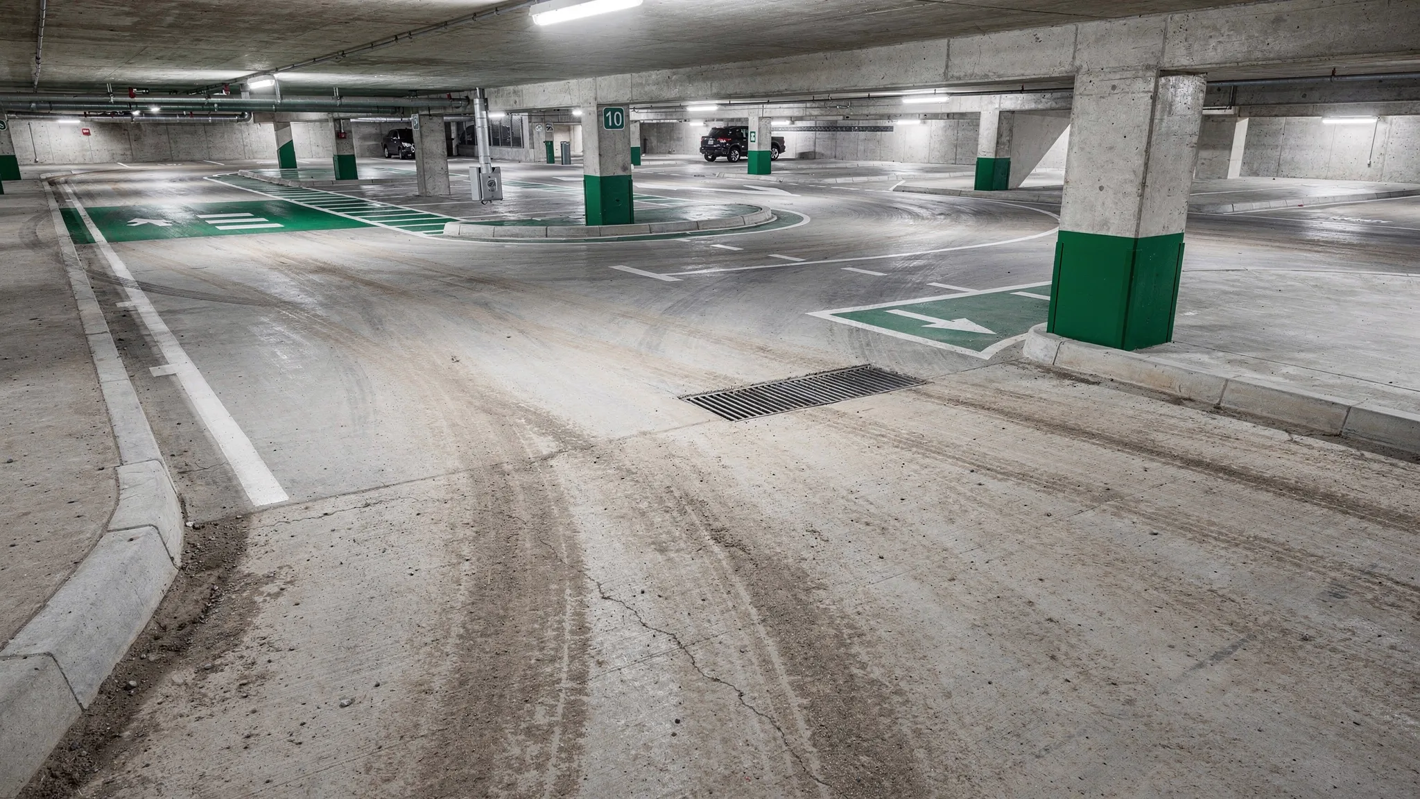 Interior of a multi-level concrete parking garage showing a ramp transition and turn lane with visible dusty tire paths, curb edges, and a floor drain area that illustrates where dust and track-out accumulate.
